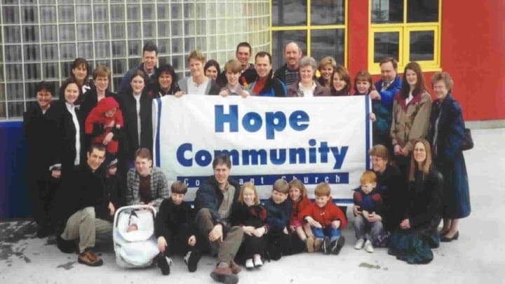 Some of the first people of Hope Community Covenant Church holding up a sign with the name of the church, outside the school they first met in.